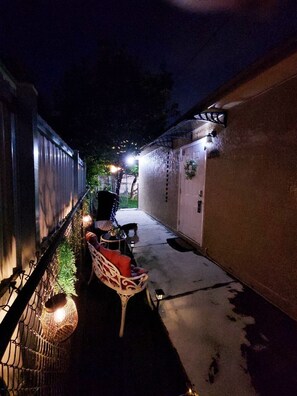 Un estrecho pasillo exterior con una silla y una mesa, una mesita con una planta, y una lámpara fijada a la pared.