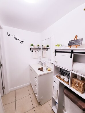 Un baño con un lavamanos blanco, un espejo, un perchero para toallas, una estantería con plantas y un cartel con las normas del baño.