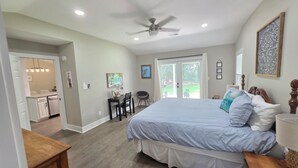 A bedroom with a bed, a ceiling fan, a desk with chairs, a dining table, and a view through a sliding door to an outdoor area. - Pelican Paradise, Piers, Pints, & Perfect Putts!  (Murrells Inlet)