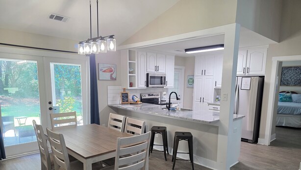 A modern kitchen with white cabinetry, a central island, and a dining area with wooden chairs. - Pelican Paradise, Piers, Pints, & Perfect Putts!  (Murrells Inlet)