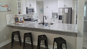 A modern kitchen with white cabinets, a stainless steel refrigerator, a microwave, and a coffee maker. There are four bar stools and a welcome sign. - Pelican Paradise, Piers, Pints, & Perfect Putts!  (Murrells Inlet)