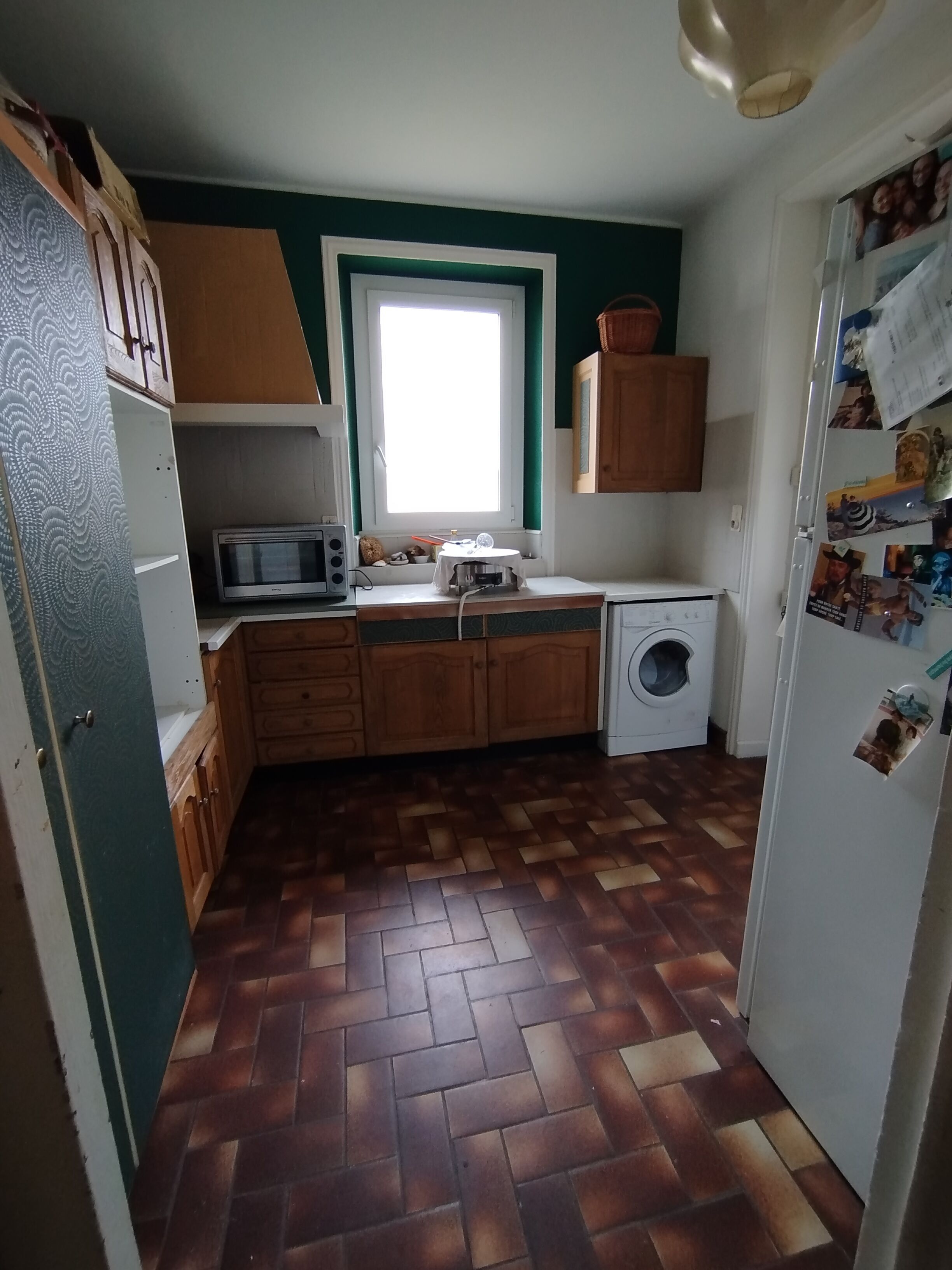 A compact kitchen with wooden cabinets, a microwave, a washing machine, and a window.