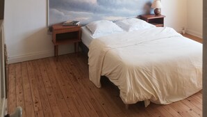 Una camera da letto con un grande murale a forma di nuvola al muro, pavimento in legno, un letto con biancheria bianca, un comodino con una lampada e una lampada a sospensione.
