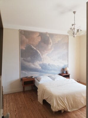 A bedroom with a large cloud mural on the wall, a wooden floor, a bed with white bedding, a nightstand with a lamp, and a hanging light fixture. - A charming, comfortable family home nestled between the sea and nature (Binic-Étables-sur-Mer)