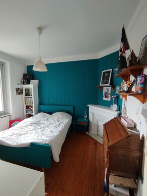 A bedroom with a teal wall, a bed with white bedding, a wooden dresser, and a shelf with various items. - Vrbo Property (Binic-Étables-sur-Mer)