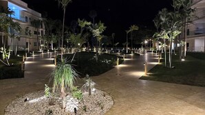 A well-lit outdoor area with a paved walkway, palm trees, and a small garden with rocks and plants. - The Surfin Turtle | Luxury Oceanfront Condo at the WAVE in Encuentro (Cabarete)
