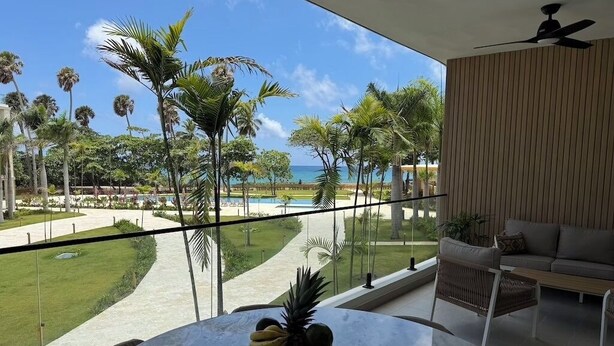 A balcony with a view of a pool and palm trees, a round table with a fruit bowl, and a modern sofa. - The Surfin Turtle | Luxury Oceanfront Condo at the WAVE in Encuentro (Cabarete)
