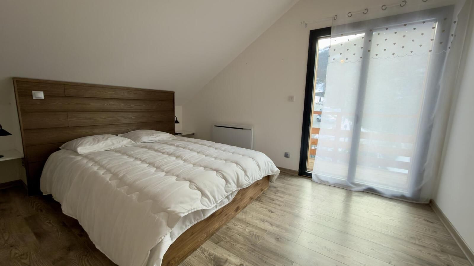 A bedroom with a wooden headboard, a large bed with white bedding, a bedside table, and a window with a curtain.