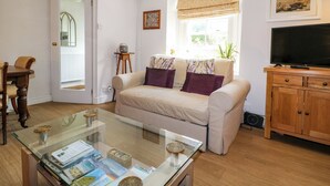 Ein Wohnzimmer mit einer Couch, einem Glastisch, einem Fernseher, einer Holzkommode und einem Fenster mit Blick ins Freie.