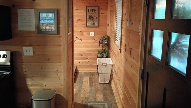 A narrow wooden cabin interior with a small kitchenette, a trash bin, a framed sign with cabin rules, and a window with blinds.