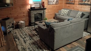 A cozy living room with a fireplace, a grey sofa, a flat-screen TV, and wooden walls.