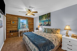 Ein Schlafzimmer mit einem Bett, einem Schreibtisch, einem Stuhl, einem Deckenventilator, einem Gemälde und einem Nachttisch mit Lampe.