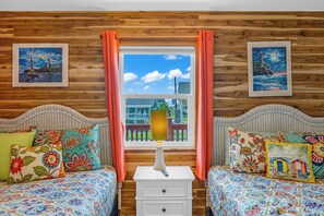 A room with two beds, wooden paneling, a window with curtains, and two framed pictures on the walls. - Crystal Beach Serenity Escape: 4BR Beachfront! Stunning Views & Outdoor Showers (Bolivar Peninsula)