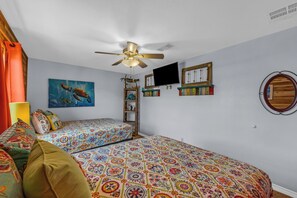A bedroom with two beds, a ceiling fan, a TV, and wall decorations including a painting and a mirror. - Crystal Beach Serenity Escape: 4BR Beachfront! Stunning Views & Outdoor Showers (Bolivar Peninsula)