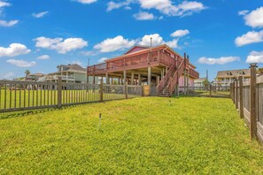 A two-story building with a balcony, surrounded by a wooden fence, set in a grassy area with clear skies. - Crystal Beach Serenity Escape: 4BR Beachfront! Stunning Views & Outdoor Showers (Bolivar Peninsula)
