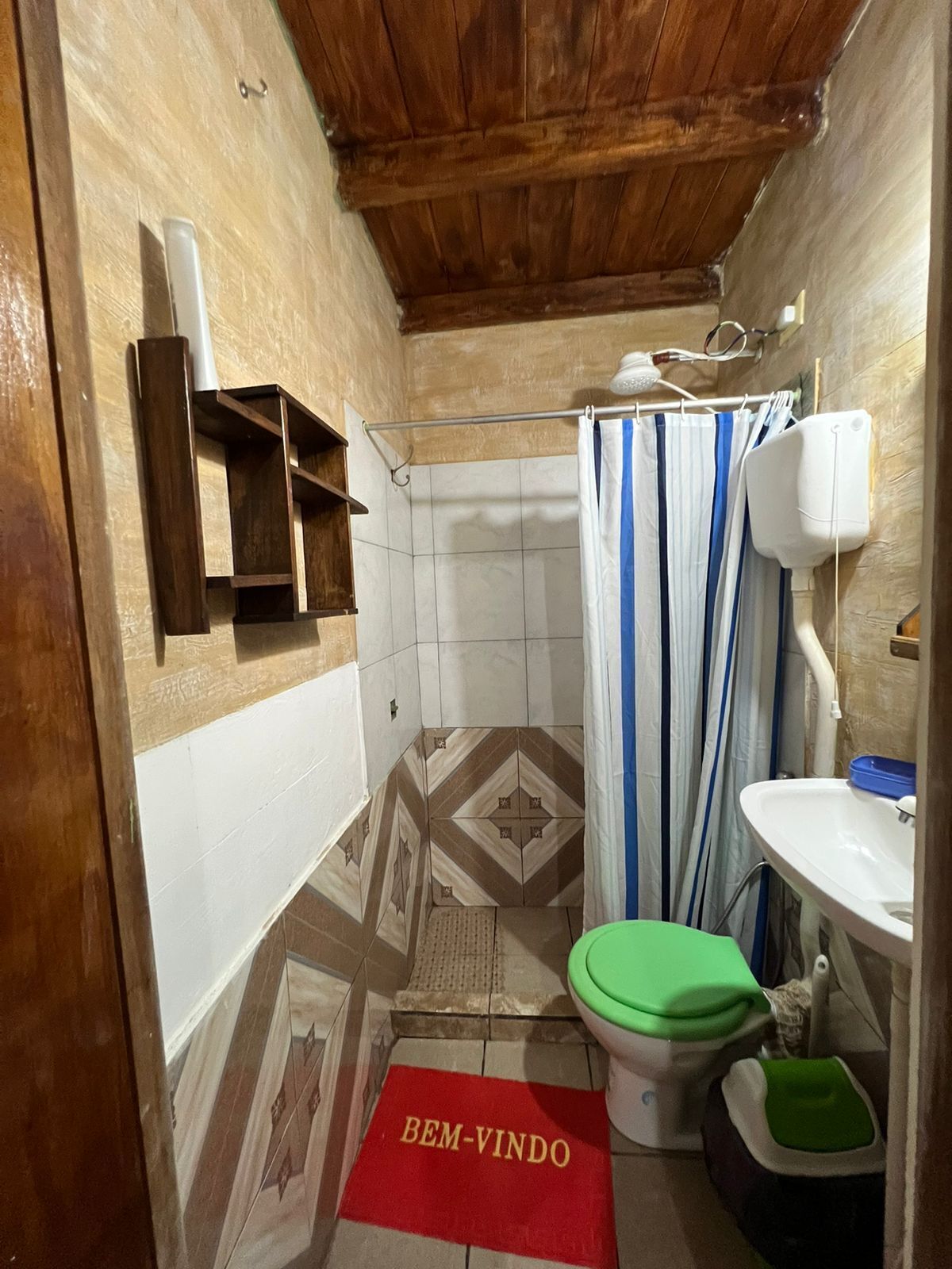 Uma casa de banho pequena com cortina de chuveiro, sanita, lavatório e prateleira com artigos de higiene pessoal.
