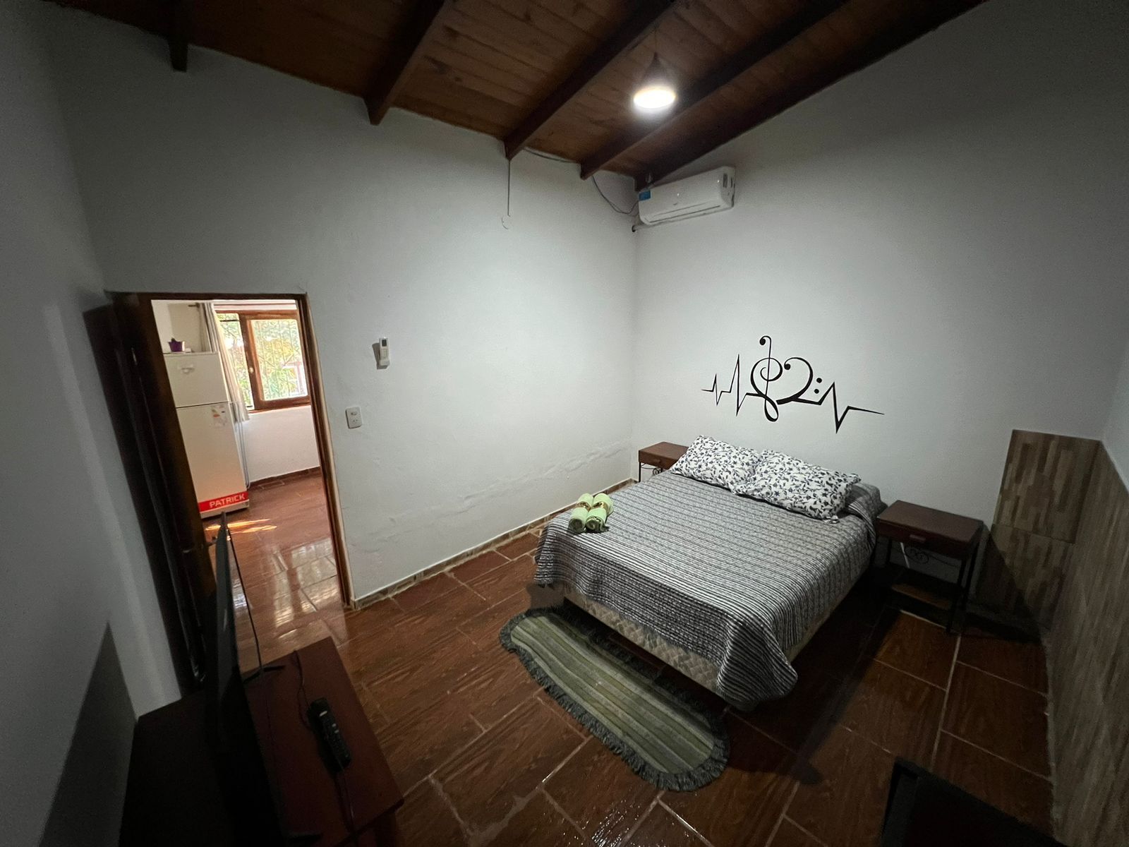 A bedroom with a bed, a nightstand, a wall-mounted air conditioner, and a wooden ceiling.