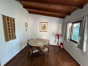 A dining area with a wooden table and chairs, a ceiling fan, a window with curtains, and a wall with a tapestry and a framed picture.