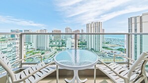 Un balcon avec une table en verre et des chaises blanches, donnant sur un paysage urbain avec de hauts immeubles.