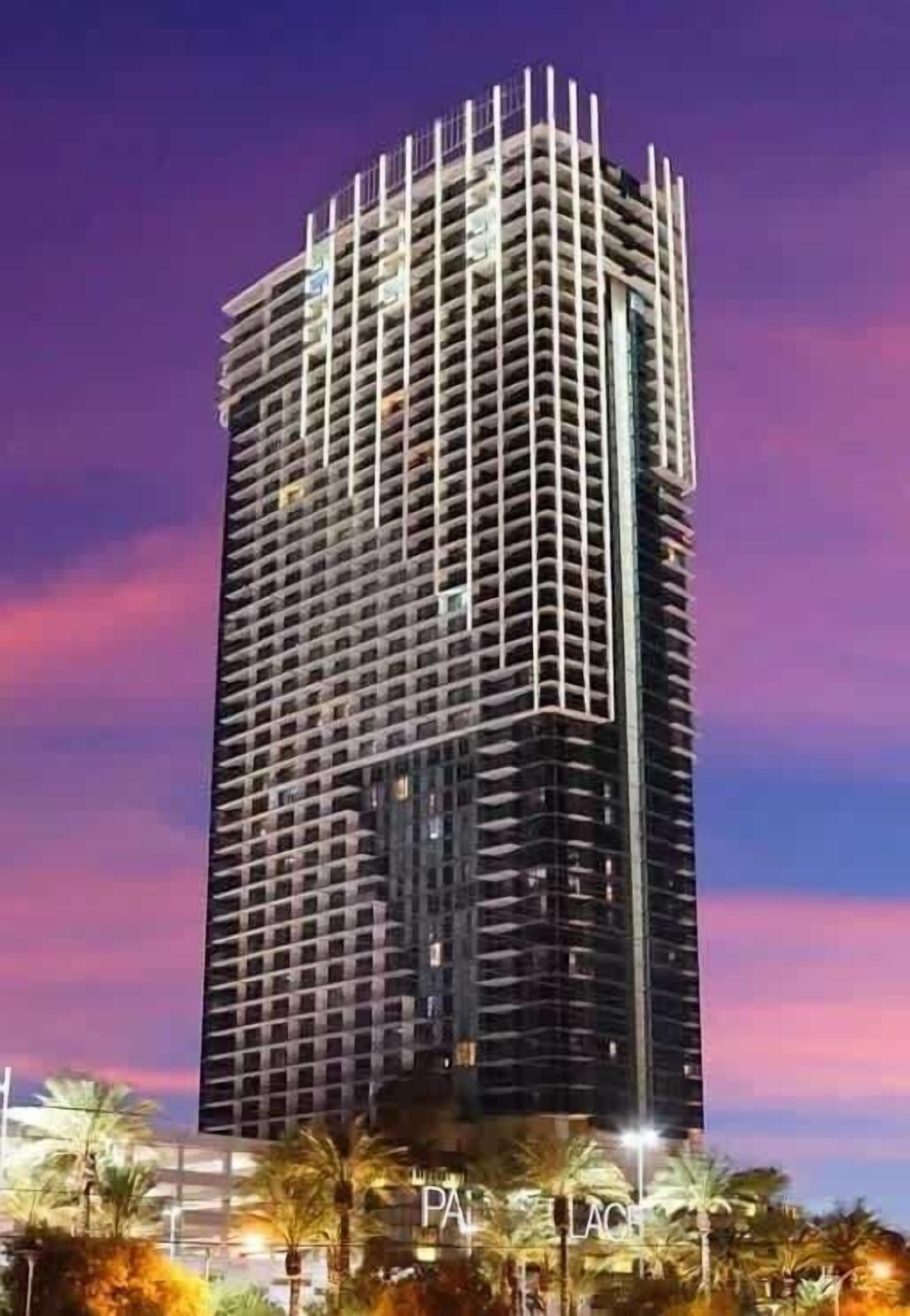A tall, modern building with a grid-like facade, surrounded by palm trees and illuminated by street lights.