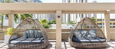 Two wicker daybeds with cushions on a rooftop terrace with a view of tall buildings.