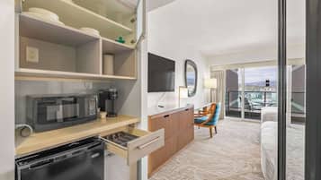A hotel room with a kitchenette, a flat-screen TV, a wooden cabinet, a chair, a bed, and a balcony with a city view.