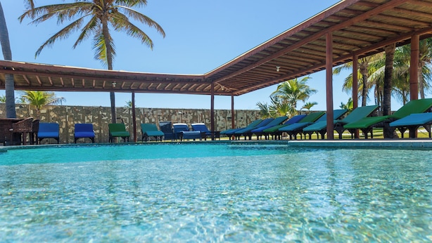 A clear pool with lounge chairs under a wooden pergola, palm trees, and a stone wall in the background.