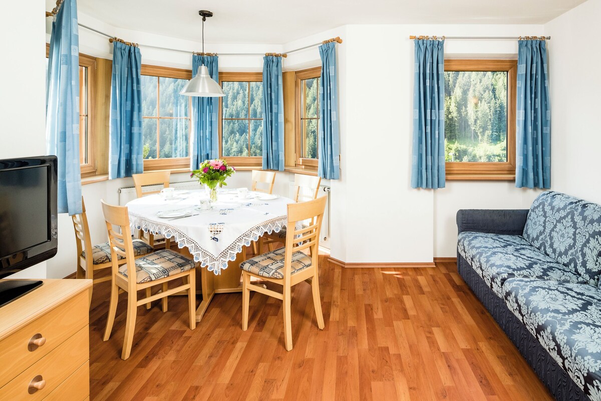 A room with a dining table set for four, a sofa, a television, and a window with curtains.