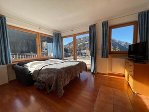 A bedroom with a large bed, a TV, a dresser, and a view of mountains through large windows. - Apartments Hetty (Santa Cristina Valgardena)