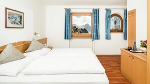 A hotel room with a bed, a nightstand, a window with curtains, a radiator, a wooden floor, a wooden wardrobe, and a framed picture on the wall. - Apartments Hetty (Santa Cristina Valgardena)