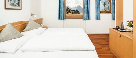 Quarto de hotel com cama, mesa de cabeceira, janela com cortinas, radiador, chão de madeira, guarda-roupa de madeira e um quadro na parede.