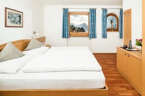 A hotel room with a bed, a nightstand, a window with curtains, a radiator, a wooden floor, a wooden wardrobe, and a framed picture on the wall. - Apartments Hetty (Santa Cristina Valgardena)