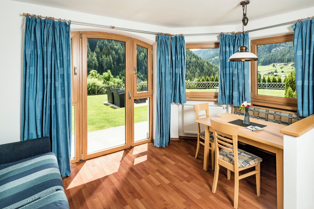 A room with a wooden floor, a dining table with chairs, a sofa, and a view of a green landscape through large windows.