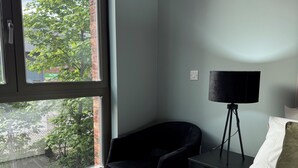 A bedroom with a large window, a black chair, a bedside table with a lamp, and a bed with white bedding. - Cozy Botanical Gardens Apartment | Sleeps 5 Guests 🌿 (Stretford)