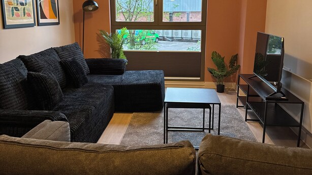 A living room with a sectional sofa, a coffee table, a TV stand, and a large window with blinds. - Cozy Botanical Gardens Apartment | Sleeps 5 Guests 🌿 (Stretford)