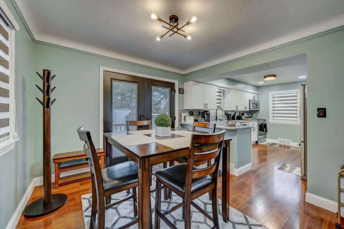 A dining area with a wooden table and chairs, a modern chandelier, and a view of the kitchen in the background.