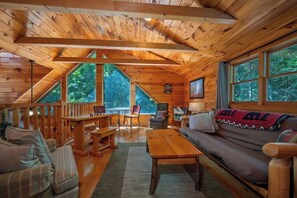 Ein Ferienhaus-Interieur mit Holzbalkendecke, großen Fenstern, einer Couch, einem Tisch und einem Sessel.