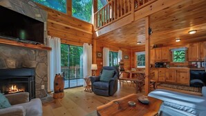 A cabin interior with a stone fireplace, wooden furniture, a dining area, and large windows. - Large log home in Lake Lure, HOT tub/Game room/pet (Lake Lure)