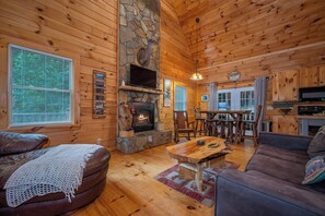 A log cabin interior with a stone fireplace, wooden furniture, and a dining area.