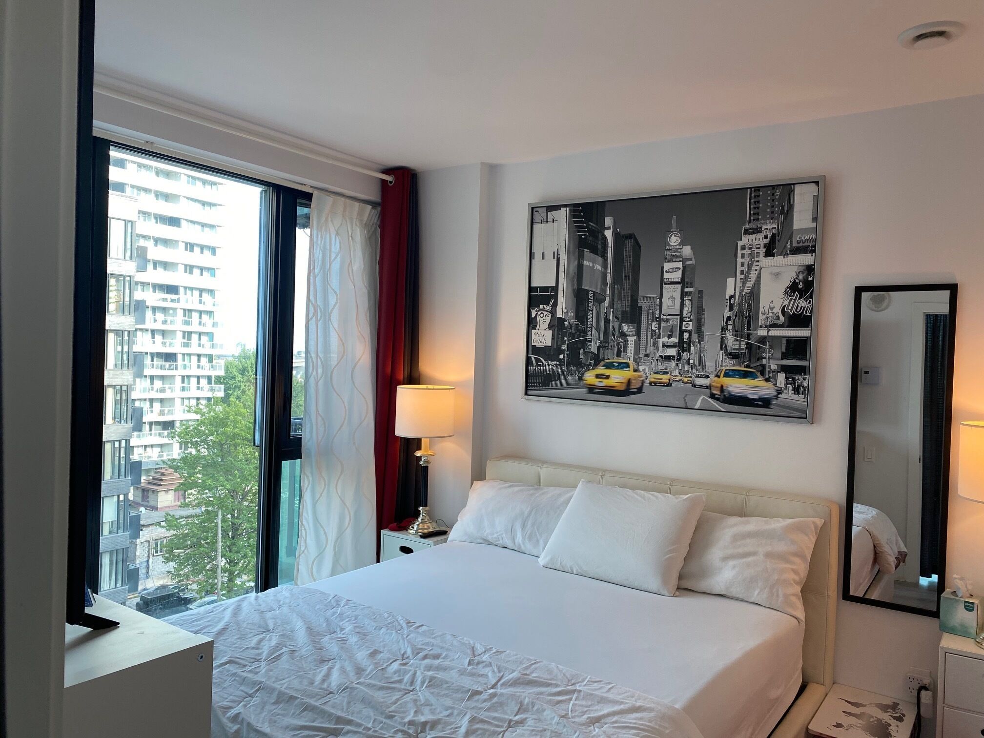 Une chambre d’hôtel avec un lit, une fenêtre avec des rideaux, une table de chevet, une lampe, un tableau accroché au mur et un miroir.