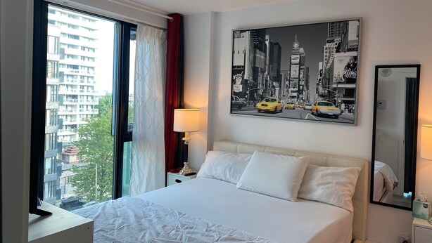 Ein Hotelzimmer mit einem Bett, einem Fenster mit Vorhängen, einem Nachttisch, einer Lampe, einem Wandbild und einem Spiegel.