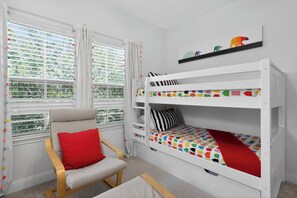Ein Kinderzimmer mit Etagenbett, einem Sessel mit rotem Kissen und einem Fenster mit Jalousien.