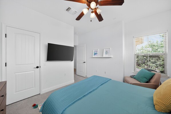A bedroom with a bed, a ceiling fan, a TV, a window with blinds, and a door.