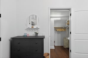 A room with a dark dresser, a mirror, and a small shelf with toiletries. There is a closet with hanging clothes and a basket on the floor.