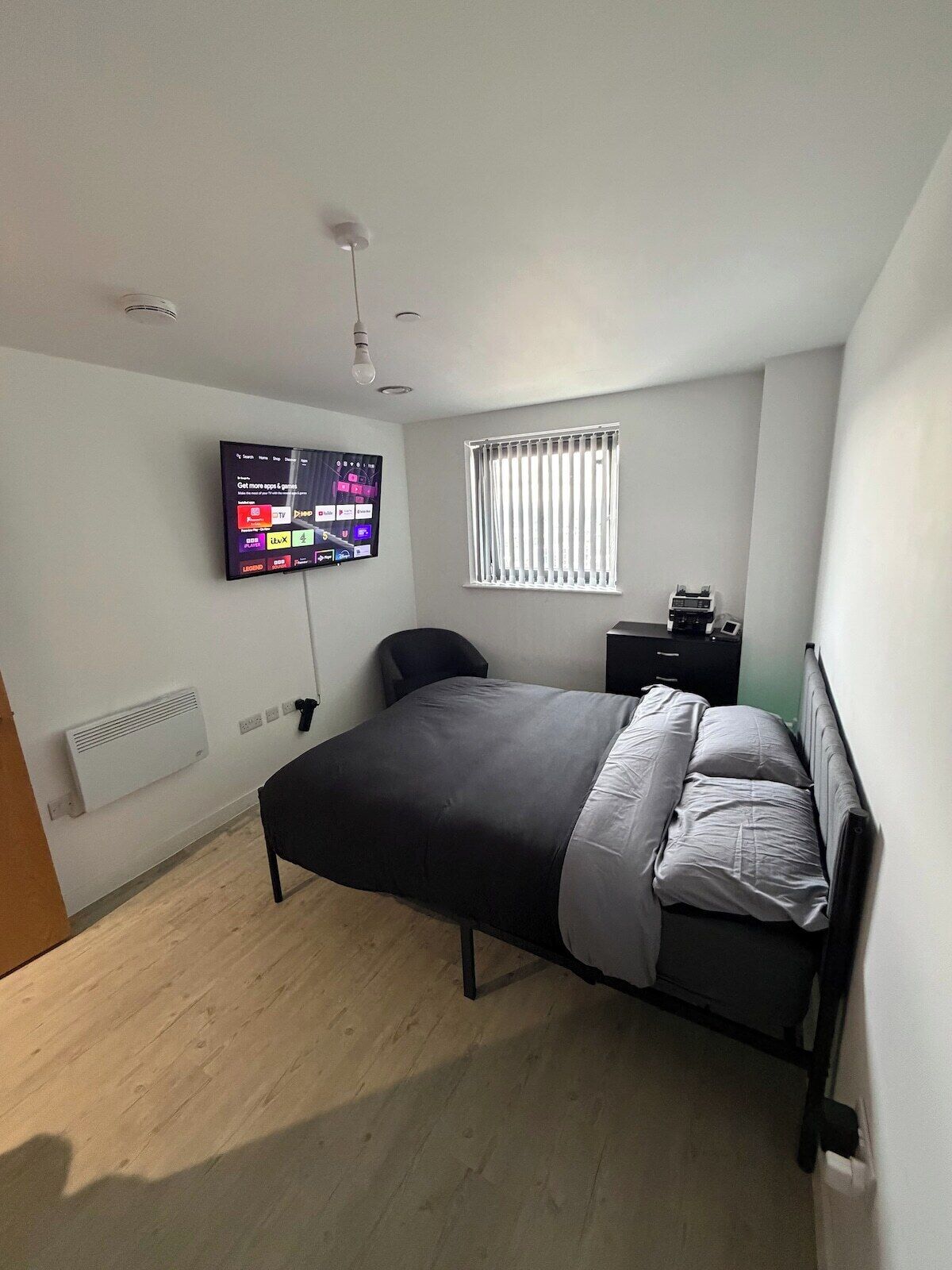 Une chambre à coucher avec un lit, un téléviseur fixé au mur, une fenêtre avec des stores et un radiateur.
