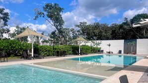 Uma piscina retangular com água cristalina, rodeada por espreguiçadeiras e uma mesa, com um toldo e vegetação ao fundo.