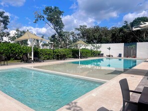 Ein rechteckiger Pool mit klarem Wasser, umgeben von Liegestühlen und einem Tisch, mit Sonnensegel und Pflanzen im Hintergrund.