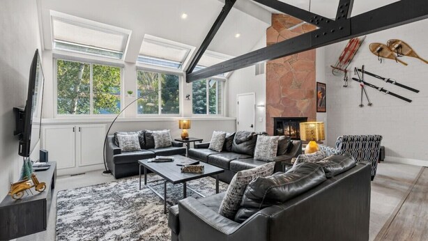 A spacious living room with a high ceiling, large windows, a fireplace, and ski equipment on the wall. - EagleVail Alpine Retreat | Pool, Golf & Pickleball (Avon)