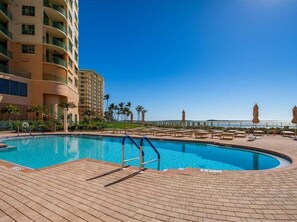 A swimming pool with a brick deck, surrounded by palm trees and a building with balconies. - Beachfront 3BR/2BA condo in Marco Island with pool, wifi, and ocean views. (Marco Island)