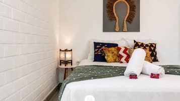 Habitación de hotel con pared de ladrillo blanco, cama con colcha verde de lunares, varios cojines de colores, una obra de arte colgada en la pared y una mesita de noche con lámpara.